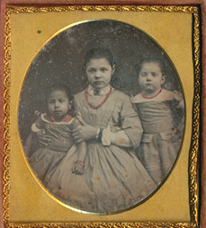 A daguerreotype with gold framing of three young girls. Photo is black and white except for red necklaces around each girls neck.